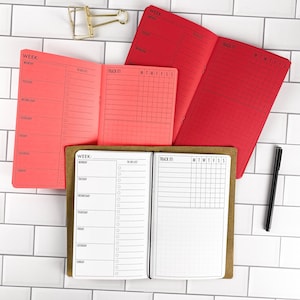 May include: Three open planners with weekly layouts and tracking grids. One planner has a brown cover with white pages, one has a coral cover with coral pages, and one has a red cover with red pages. A gold binder clip and a black pen are included.
