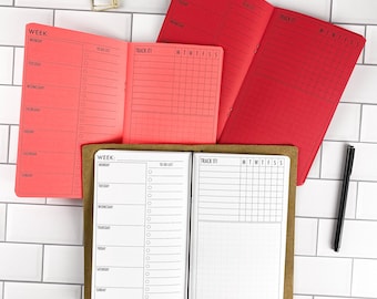 May include: Three open planners with weekly layouts and tracking grids. One planner has a brown cover with white pages, one has a coral cover with coral pages, and one has a red cover with red pages. A gold binder clip and a black pen are included.