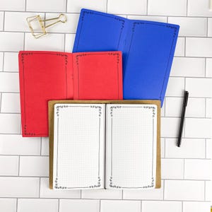 May include: Three open notebooks with dot grid paper and black floral border designs. One notebook is blue, one is red, and one is white with a brown cover. A gold metal binder clip and a black pen are positioned near the notebooks.