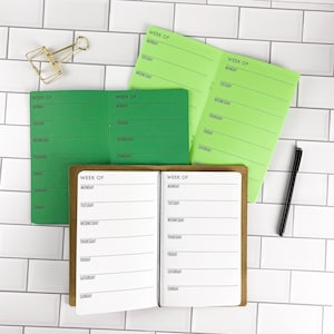 May include: Three weekly planners with black text on white, green, and lime green paper. Each planner features a header reading WEEK OF followed by a list of days from Monday to Sunday. A gold metal binder clip and a black pen are included.