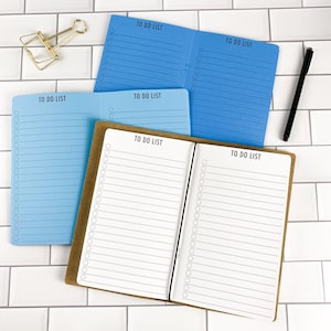 May include: Three open to-do list notebooks in blue, light blue, and white with brown covers. Each page features horizontal lines and circular checkboxes. A gold metal binder clip and a black pen are positioned near the notebooks.