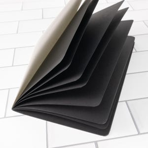 May include: A notebook with a light brown cover and multiple internal pages made of solid black paper. The notebook is partially open, displaying the contrast between the tan exterior and the dark interior sheets, with rounded corners on the pages.