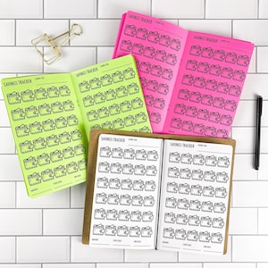 May include: Three open savings tracker booklets in neon green, bright pink, and white with brown covers. Each page features rows of black wallet icons with dollar signs, along with printed fields for savings goals, amounts, start dates, and end dates.