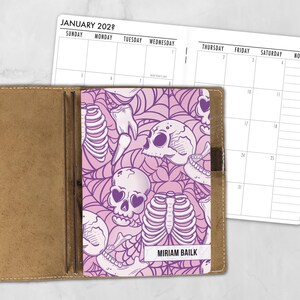 May include: A planner insert featuring a purple and light pink pattern of skulls, rib cages, and spiderwebs. The cover includes the text MIRIAM BAILK. The open calendar pages show the month of JANUARY 202? with a grid layout for daily scheduling.