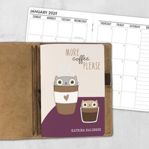 May include: A tan leather notebook cover holding a planner insert with a calendar page visible. The notebook cover features a graphic of two cats in coffee cups with the text "MORE coffee PLEASE" and "KATRINA BALINESE" printed on the front.