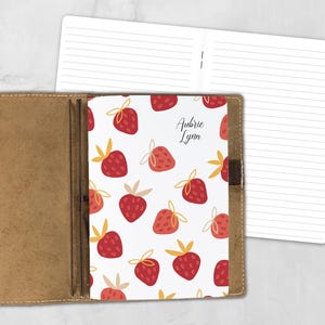 May include: A tan leather notebook cover holding a white notebook insert with a repeating red strawberry pattern and yellow leaves. The name Aubrie Lynn is printed in black script on the cover. A second open notebook with lined pages is visible.