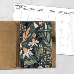 May include: A tan leather planner cover holding a notebook with a dark green botanical cover featuring orange and white flowers, leaves, and berries. The name Madison Jungson is printed on the cover. A January calendar page is visible behind it.