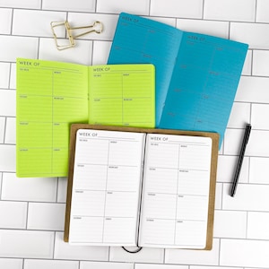May include: Three open weekly planners in bright lime green, vibrant blue, and white with brown covers. Each planner features a grid layout with the text "WEEK OF" at the top and days of the week listed. A gold metal binder clip and a black pen are included.