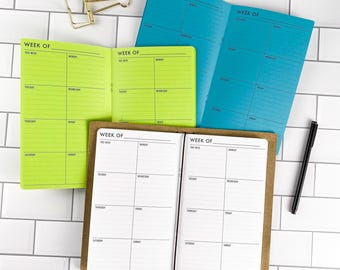 May include: Three open weekly planners in bright lime green, vibrant blue, and white with brown covers. Each planner features a grid layout with the text "WEEK OF" at the top and days of the week listed. A gold metal binder clip and a black pen are included.