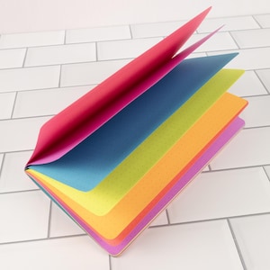 May include: A multi-colored notebook with a softcover featuring pages in vibrant shades of pink, blue, lime green, orange, and purple. Each page is printed with a subtle dot grid pattern, and the notebook is partially open to display the layered colors.