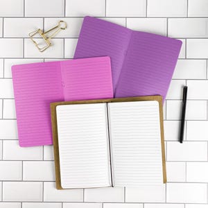 May include: Three open notebooks with lined pages in purple, pink, and brown kraft paper covers. A gold-toned metal binder clip is positioned in the upper left corner, and a black ballpoint pen lies vertically on the right side of the notebooks.