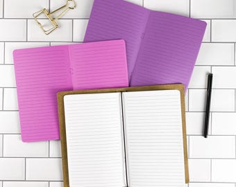May include: Three open notebooks with lined pages in purple, pink, and brown kraft paper covers. A gold-toned metal binder clip is positioned in the upper left corner, and a black ballpoint pen lies vertically on the right side of the notebooks.