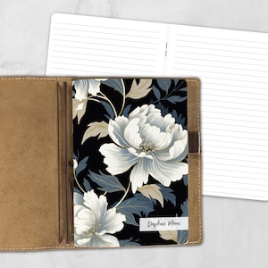 May include: A notebook with a black cover featuring a white and blue floral pattern and the text Daphne Moon. The notebook is held within a tan leather-like cover with elastic bands, and an open lined notebook page is visible behind it.