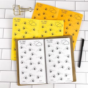 May include: Three open savings challenge booklets with yellow and white pages featuring bee illustrations, sun and cloud icons, and the text $100 SAVINGS CHALLENGE. A gold metal binder clip and a black pen are positioned near the booklets.