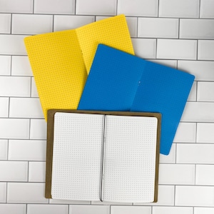 May include: Three open notebooks with dot grid paper. One notebook has a bright yellow cover, one has a vibrant blue cover, and the third has a brown kraft paper cover with a black elastic band holding the white pages open in the center.