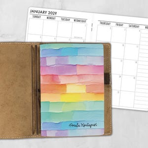 May include: A tan leather planner cover holding a notebook with a colorful, textured, horizontal brushstroke pattern in pastel shades of blue, pink, yellow, and green. The notebook cover features the text Amala Koutapuri and an open calendar page.