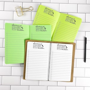 May include: Three open notebooks with lime green or white pages featuring black printed lines, checkboxes, and the text "obsessive LIST maker" with heart icons. A gold metal binder clip and a black pen are positioned near the notebooks.