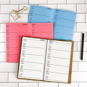 May include: Three open contact address books in light blue, bright pink, and brown with white pages featuring black printed lines and the text "CONTACTS" at the top of each page. A gold metal binder clip and a black pen are positioned nearby.