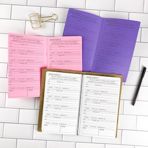 May include: Three open prayer request journals in pink, purple, and white, each featuring lined sections for date, prayer for, and need, with checkboxes for urgent, fulfilled, and answered on. A gold metal binder clip and a black pen are included.