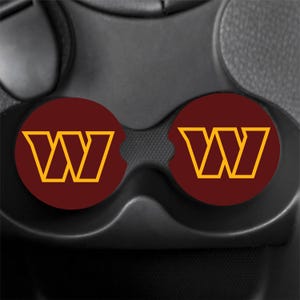 May include: Two circular car cup holder inserts featuring a deep burgundy background with a prominent gold-coloured letter W logo centred on each. These round coasters are designed to fit into standard vehicle console cup holders for protection.