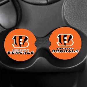 Op de afbeelding: Twee ronde bekerhouder-onderzetters voor in de auto met het oranje en zwarte logo van de Cincinnati Bengals en de teamnaam in zwarte letters eronder gedrukt. De onderzetters zijn geplaatst in een zwarte bekerhouderconsole van een voertuig.