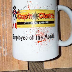 May include: A white ceramic mug featuring a graphic of a pirate holding two swords, a yellow rectangular sign with the text Cap'n Clark's OTTOMAN EMPIRE, and the text Employee of the Month, all accented by red paint splatter designs.