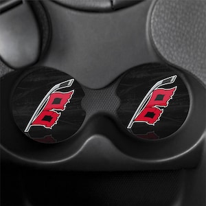 May include: Two circular car cup holder inserts featuring a black background with a distressed texture. Each insert displays a graphic of a red flag with a black square center, attached to a white and grey hockey stick handle design.