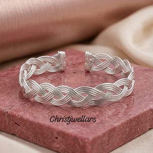 May include: A silver-toned cuff bracelet featuring a multi-strand braided wire design with an open back. The bracelet has a polished metallic finish and is displayed with the text Christjwellars printed in black on the surface below it.