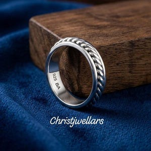 Puede incluir: Un anillo de plata con un patrón de cuerda trenzada en la banda exterior. El interior de la banda está grabado con el texto 925 BA. La palabra Christjwellars aparece en texto blanco debajo del anillo. El anillo tiene un acabado metálico pulido.