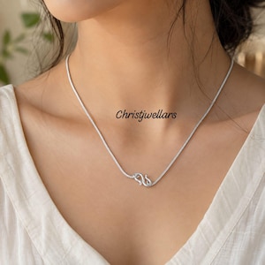 Può includere: Una collana a catena color argento con un piccolo dettaglio di chiusura curvo a forma di S posizionato sul davanti. Il metallo ha una finitura lucida e il testo Christjwellars è visibile in lettere nere vicino al centro della catena.