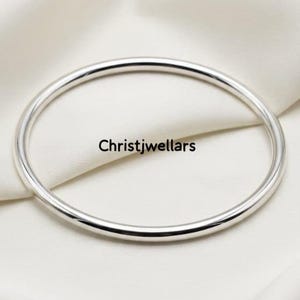 Peut inclure: Un bracelet jonc en argent poli avec une surface lisse et arrondie. Le texte Christjwellars est imprimé en noir au centre de l'ouverture circulaire du bracelet en argent, qui présente une finition métallique réfléchissante et un design simple.