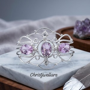 Può includere: Un bracciale rigido in argento con tre pietre preziose di ametista color viola chiaro dal taglio ovale, incastonate in una complessa montatura in metallo traforato. La pietra centrale è affiancata da due pietre più piccole e il testo Christjwellars.