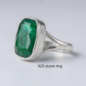 Pode incluir: Um anel 925 stone ring com uma grande pedra verde retangular facetada montada numa banda de prata polida. A banda de metal tem um design de haste dividida que se liga à cravação da pedra verde. O texto 925 stone ring é visível.
