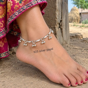 May include: A 925 silver payal anklet featuring a delicate chain with small, dangling spherical bells attached at regular intervals. The metallic silver links are connected by circular rings, creating a flexible band that rests around the ankle.