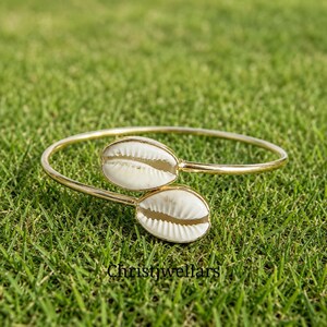 May include: A gold-toned open cuff bracelet featuring two white cowrie shells at the ends. The metal band is thin and polished, holding the shells securely in place. The text Christjwellars is visible at the bottom of the image.