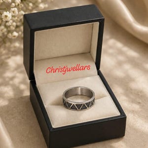 May include: A silver-toned metal ring featuring a repeating black triangular pattern around the band, presented inside a black jewellery box with a cream-coloured interior. The text Christjwellars is printed in red on the inner lid of the box.