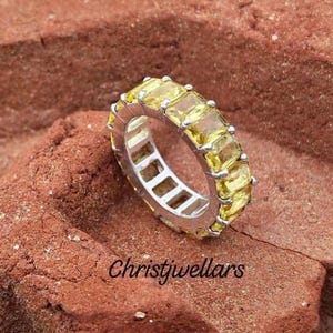Può includere: Un anello eternity in argento con una fila continua di pietre preziose rettangolari gialle sfaccettate montate a griffe. L'anello è posizionato su una superficie ruvida color marrone rossastro. Il testo Christjwellars è visibile sotto l'anello.