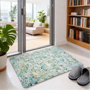 May include: A rectangular floor mat featuring a repeating pattern of smooth, rounded river stones in various shades of light blue, teal, sage green, and cream. The mat lies flat on a light-colored floor, positioned near a pair of black and white checkered slippers.