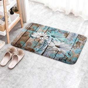 May include: A rectangular floor mat featuring a rustic wood plank pattern in shades of brown and distressed teal. The design includes large white and light blue daisy-like flowers with dark centers, accented by small black butterfly silhouettes.