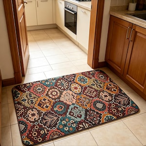 May include: A rectangular floor mat featuring a multi-colored patchwork pattern with various geometric, floral, and abstract shapes in shades of deep red, teal, orange, brown, and cream, finished with a dark brown border on a tiled kitchen floor.