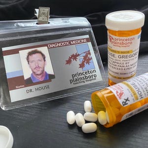 Op de afbeelding: Een doorzichtige plastic badgehouder met een kaart van het Princeton Plainsboro Teaching Hospital voor Dr. House, naast twee oranje medicijnflesjes met witte doppen en verschillende witte ovale pillen verspreid over het oppervlak.