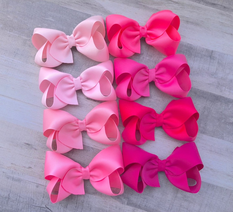 May include: Nine pink hair bows in varying shades of pink, from light to deep fuchsia. The bows are made of a smooth, satin-like material and are arranged in three rows of three.