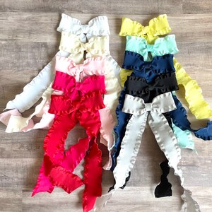 Pu&ograve; includere: Un set di dieci fiocchi di tessuto arricciati in vari colori, tra cui bianco, giallo, azzurro, blu scuro, rosa e rosso. I fiocchi sono disposti in una pila, con il fiocco pi&ugrave; grande in basso e il fiocco pi&ugrave; piccolo in alto.