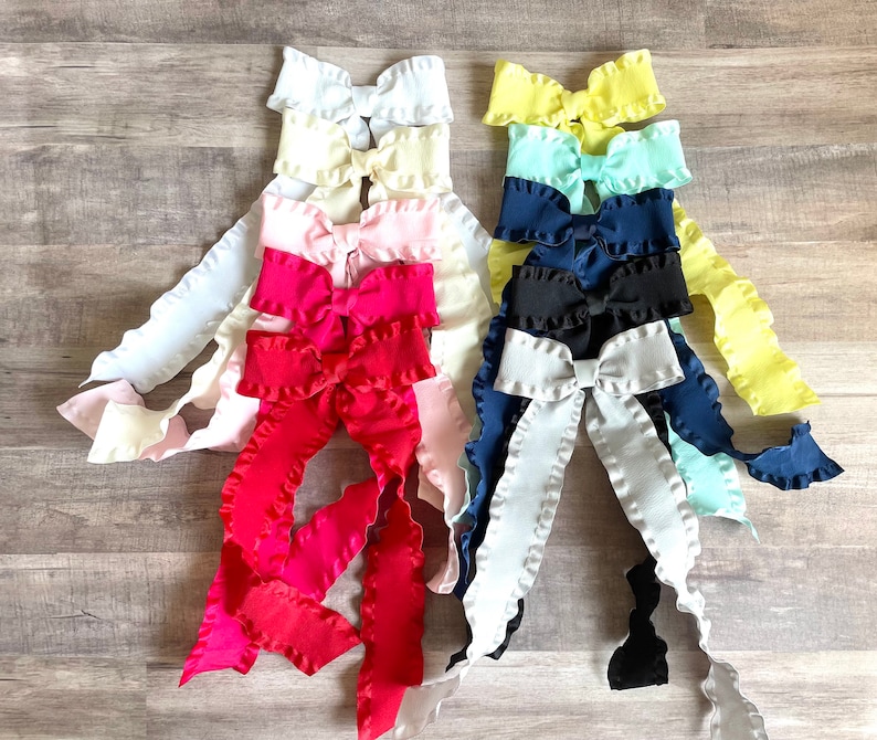 Pu&ograve; includere: Un set di dieci fiocchi di tessuto arricciati in vari colori, tra cui bianco, giallo, azzurro, blu scuro, rosa e rosso. I fiocchi sono disposti in una pila, con il fiocco pi&ugrave; grande in basso e il fiocco pi&ugrave; piccolo in alto.