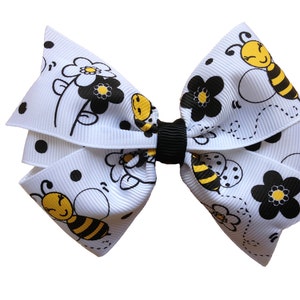 May include: White hair bow with black polka dots and a bee design. The bee is yellow and black with a smiling face. The bow has black and white flowers with yellow centres.