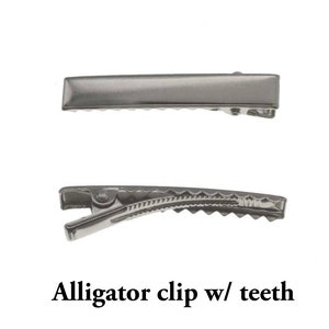 K&ouml;nnte beinhalten: Zwei silberfarbene Haarspangen vom Typ Alligator vor wei&szlig;em Hintergrund. Die obere Spange ist rechteckig, w&auml;hrend die untere Z&auml;hne hat. Der Text "Alligator clip w/ teeth" steht darunter.