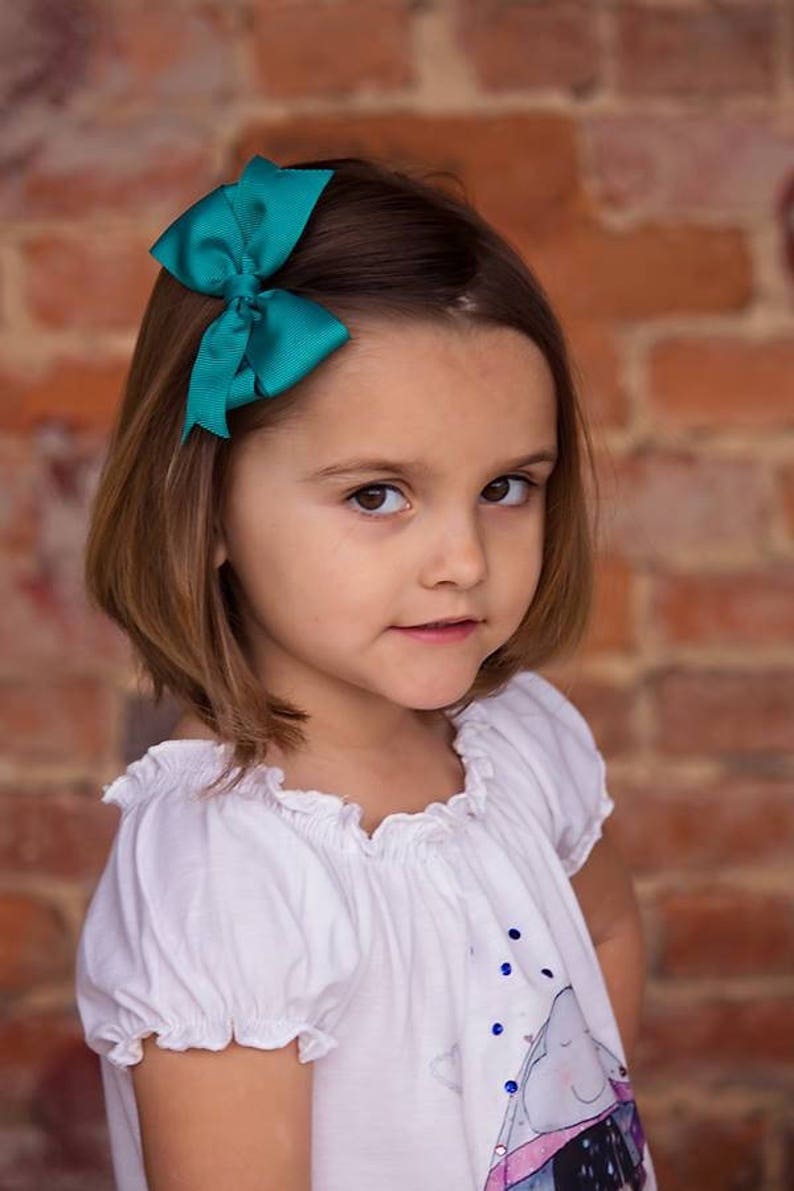 Pode incluir: Uma menina jovem com cabelo castanho curto vestindo uma camisa branca com uma estampa gr&aacute;fica azul e branca. Ela est&aacute; usando um la&ccedil;o turquesa no cabelo.
