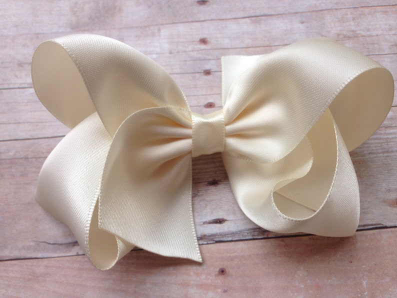 May include: A large ivory satin bow with a simple center knot. The bow is made from a soft, luxurious fabric and is perfect for adding a touch of elegance to any outfit.
