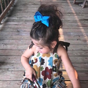 May include: A young girl with long brown hair wearing a floral print dress with a blue bow in her hair. She is sitting on a wooden deck.