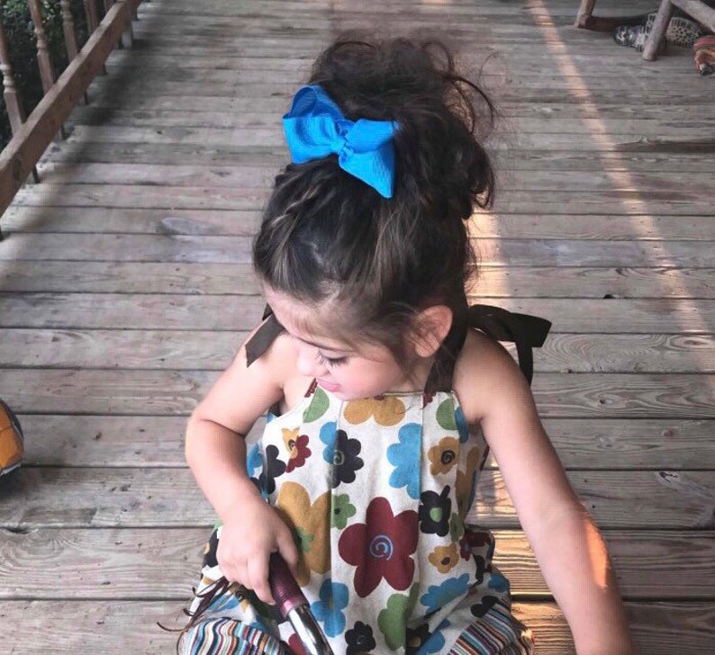 May include: A young girl with long brown hair wearing a floral print dress with a blue bow in her hair. She is sitting on a wooden deck.